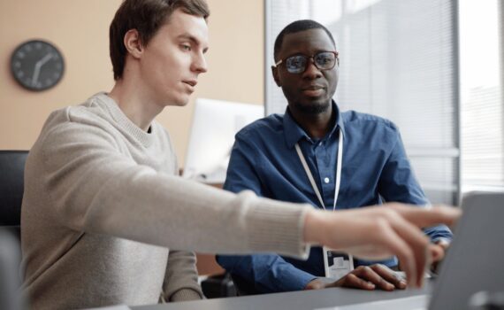 Two colleagues are looking at a laptop screen in an office setting, with one pointing at the screen, illustrating a moment of instruction or review relevant to cybersecurity training.
